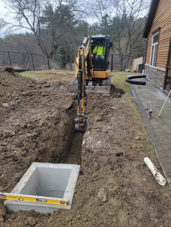 Excavated drainage trench with perforated pipe and gravel backfill installed along the side of a residential property in Lansing, Michigan. The surrounding yard shows clay-heavy soil typical of mid-Michigan, with the drainage system routed away from the home's foundation.