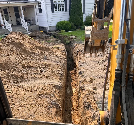 An open utility trench on a Lansing residential property showing a newly installed sewer lateral with compacted backfill alongside the pipe. A TMT Excavation excavator sits on the job site with the trench extending toward the street connection point.